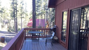 Une terrasse en bois avec une table et des chaises, une porte vitrée et une vue sur une zone boisée.