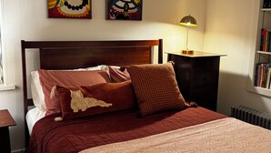 A neatly made bed with a red and white patterned bedspread, a brown pillow, and a decorative pillow with a cat design. There is a wooden nightstand with a lamp and a bookshelf with books. Two paintings are hung on the wall above the bed. - Beautiful Large Loft on Historic Water Street in Downtown Decorah (Decorah)