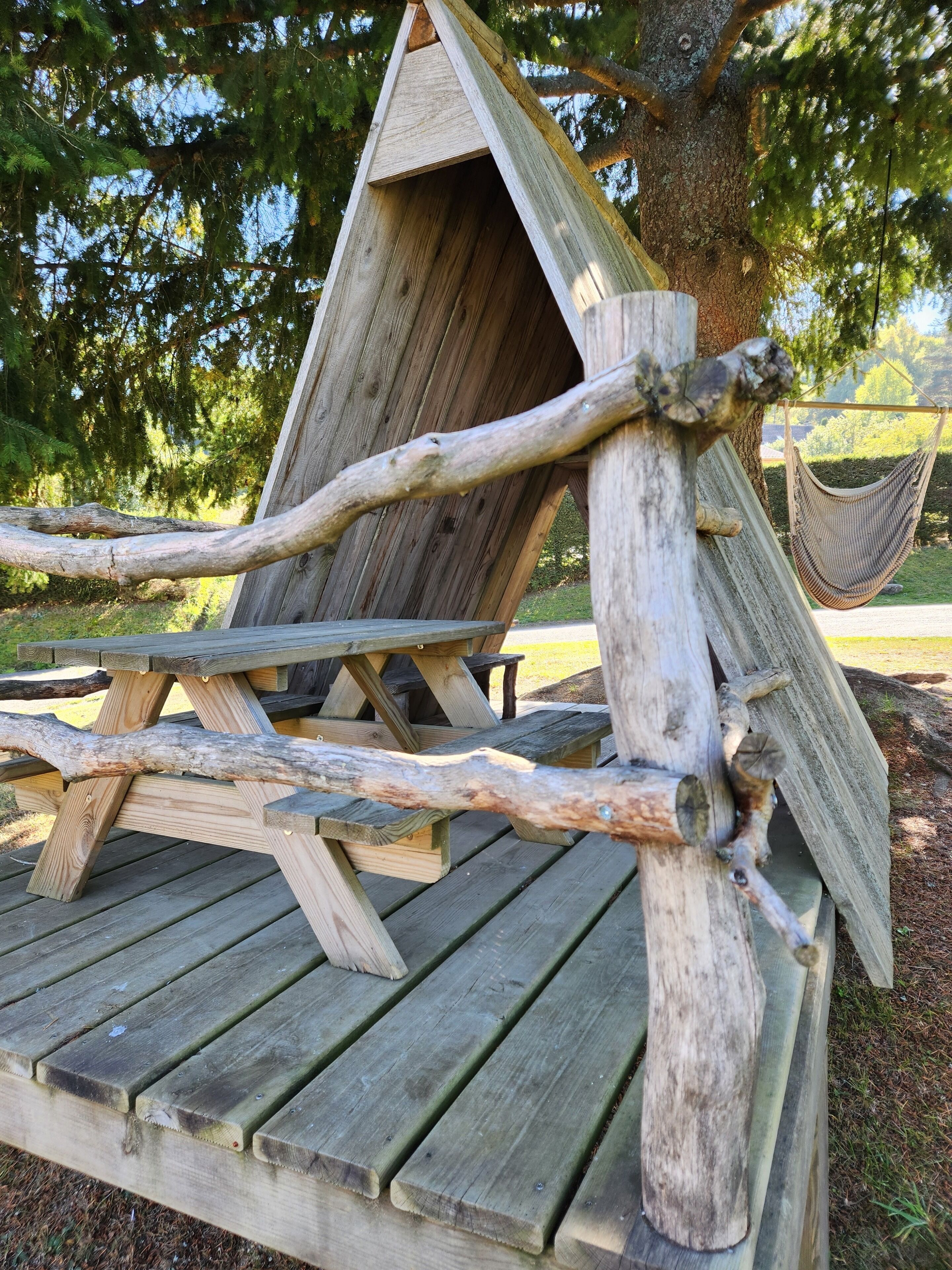 Ein Holztisch für ein Picknick mit einer Hängematte und einer Holzkonstruktion, die einem Vogelhaus oder einer Laube ähnelt.