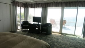 Ein Hotelzimmer mit einem großen Bett, einem Fernseher auf einem Schrank, einem Schreibtisch und Meerblick durch große Fenster.