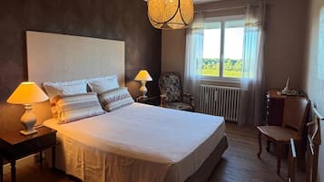 Ett sovrum med en stor säng, två sänglampor, en stol, ett skrivbord och ett fönster med utsikt över grönområden.