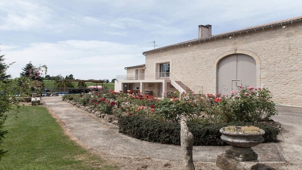 A stone building with a large garage door, a covered outdoor area with a pergola, and a garden with pink flowers.