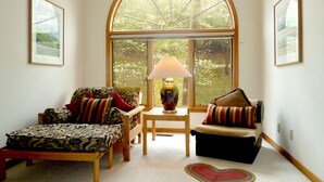 Una habitación acogedora con un gran ventanal arqueado, una cama, una silla, una mesa con lámpara y un tapete con forma de corazón.