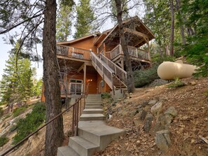 Une cabane en bois à plusieurs niveaux, avec un escalier menant à un balcon, entourée d’arbres et située sur une colline rocheuse.