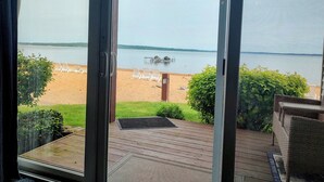 Vue depuis une chambre dotée d’une grande porte vitrée donnant sur une terrasse surplombant une plage, avec des chaises longues et un quai.