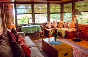 Um quarto acolhedor com um sofá, uma mesa de centro de madeira com um vaso, uma mesa de jantar com toalha verde e um banco com almofadas estampadas.