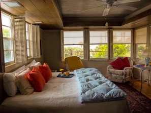 Quarto com cama, poltrona, mesa e janela com vista para o verde.