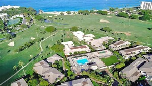 An aerial view of a golf course with a swimming pool, surrounded by residential buildings and a beach.