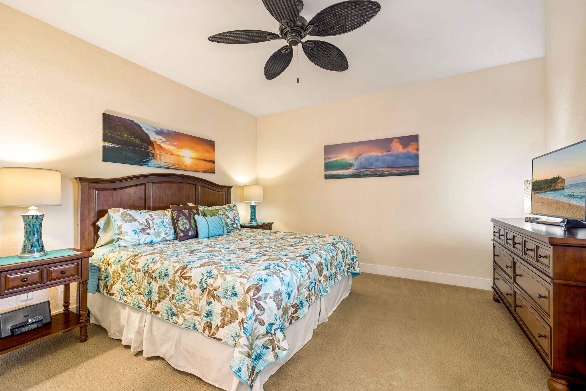 Une chambre à coucher comprenant un lit, deux tables de chevet, une commode, un ventilateur de plafond et deux tableaux accrochés au mur.