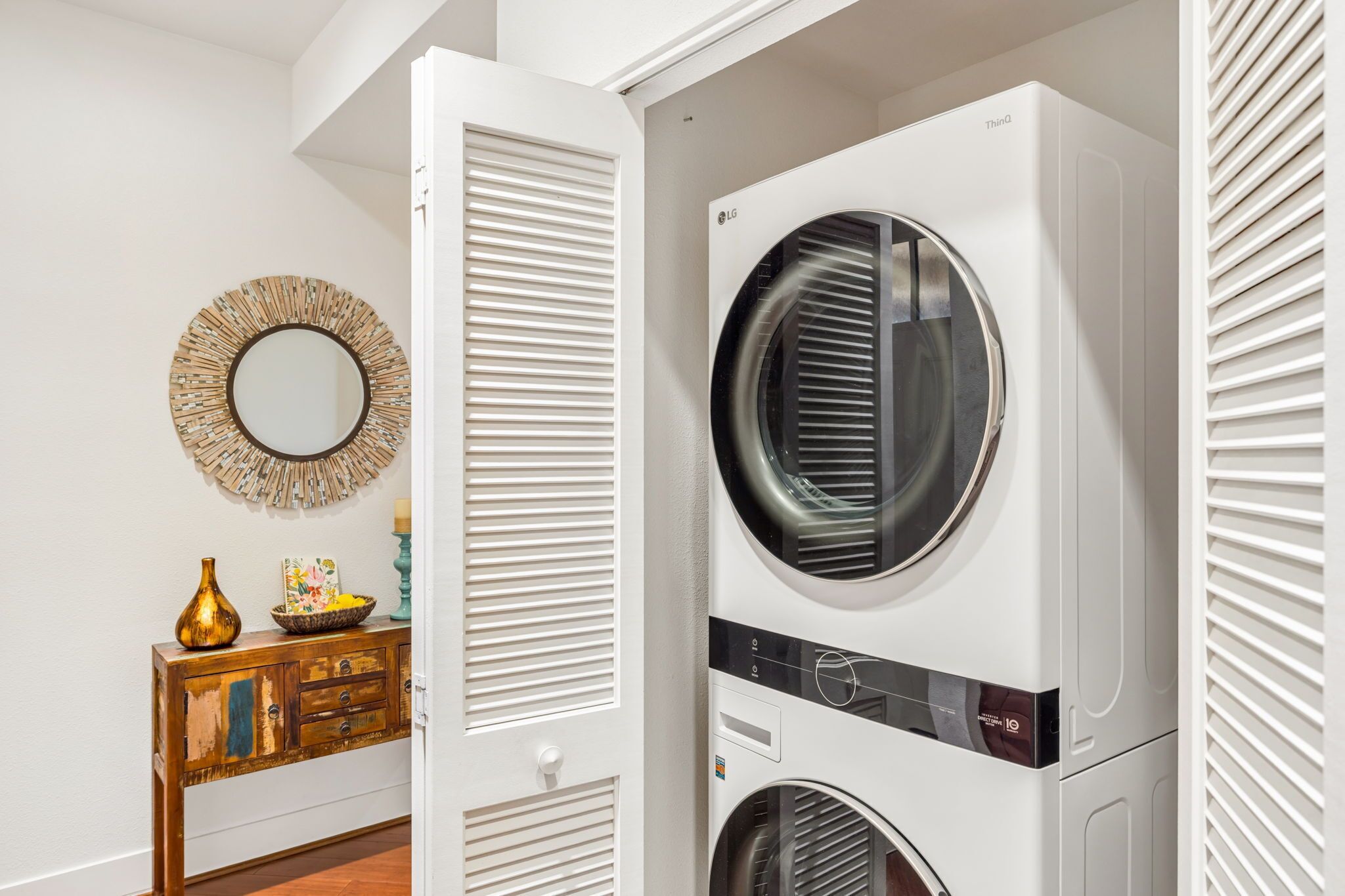 A laundry room with a stacked washer and dryer, a decorative mirror, and a wooden dresser with a vase and picture frame.