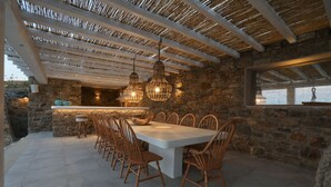 A dining area with a stone wall backdrop, a large table, and hanging light fixtures. - Villa Moonflower (Mykonos)