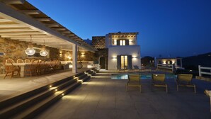 A modern outdoor dining area with a pool, stone walls, and a two-story building with illuminated windows. - Villa Moonflower (Mykonos)