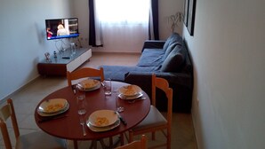 Ein Essbereich mit Tisch für zwei Personen, einer Couch, einem Fernseher und einem Fenster mit Vorhängen.