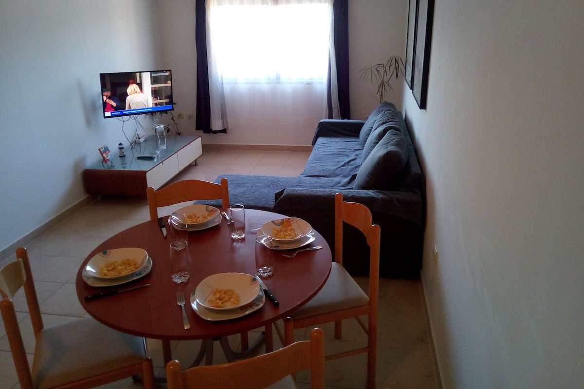 Ein Essbereich mit Tisch für zwei Personen, einer Couch, einem Fernseher und einem Fenster mit Vorhängen.