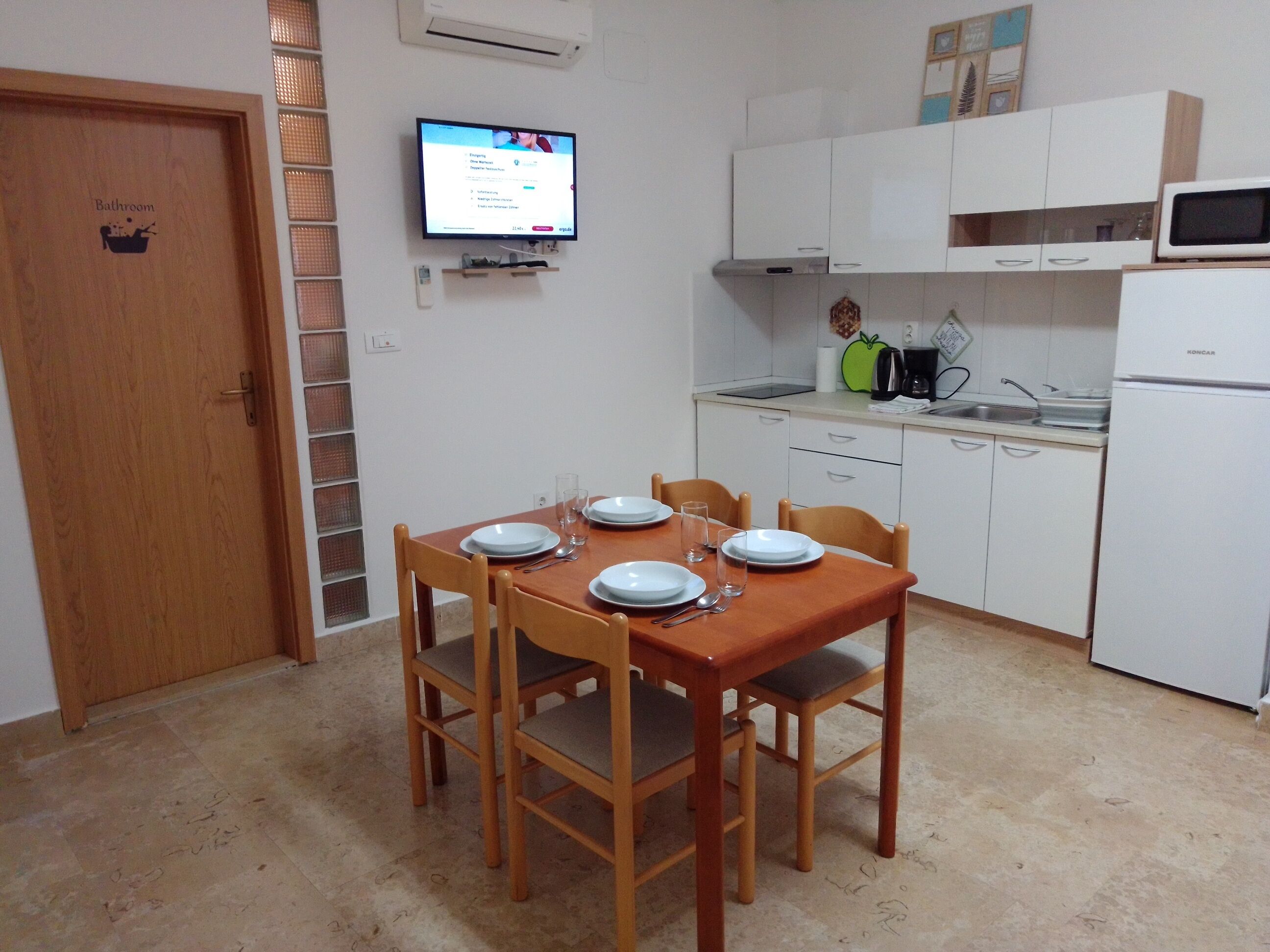 A small, clean kitchen with white cabinets, a microwave, and a sink. There is a wooden dining table with four chairs and plates set for a meal. A television is mounted on the wall above the table.