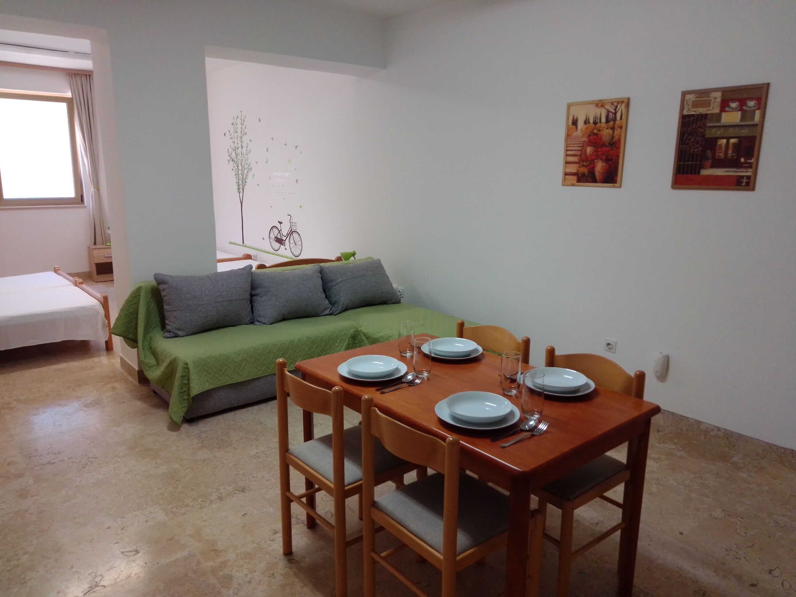 A dining area with a wooden table set for four, a green sofa, and a small bed in the background.