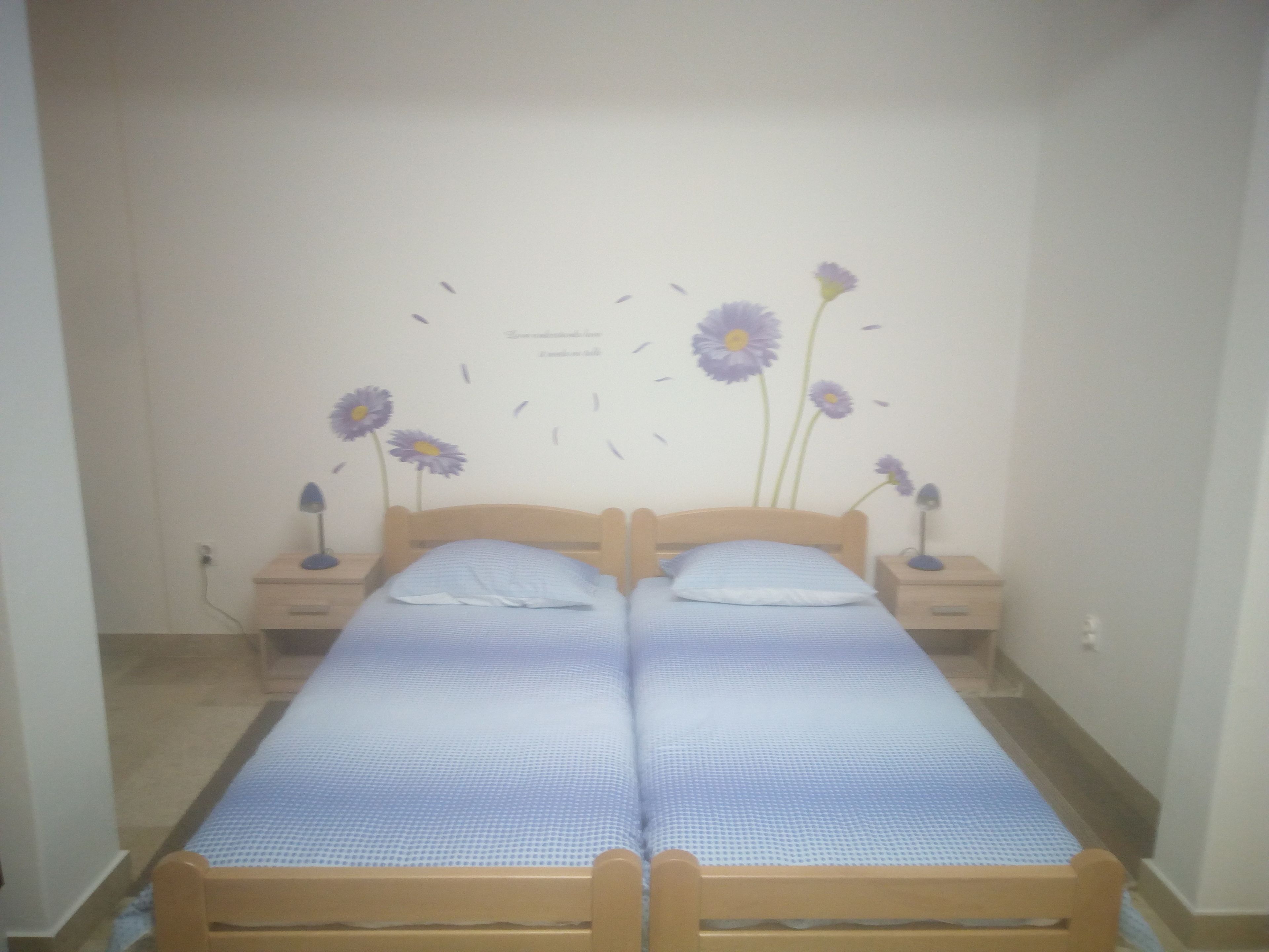 Uma cama de casal com colcha xadrez azul, duas mesas de cabeceira com abajures e adesivos de flores roxas na parede.