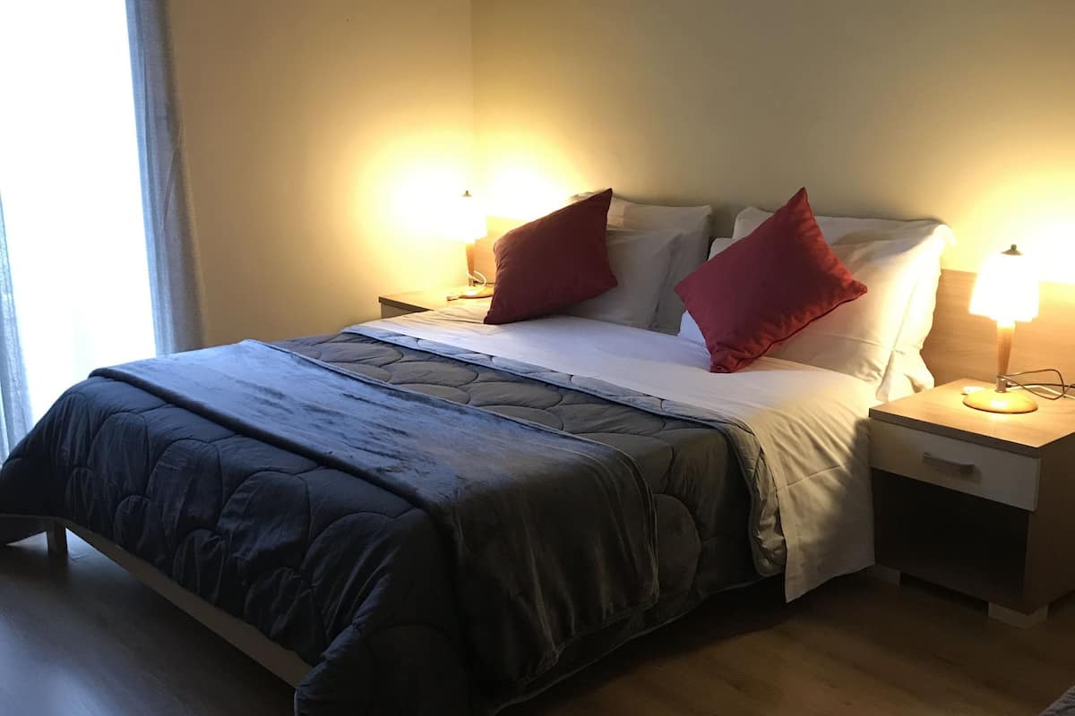 A hotel room with a bed, two bedside tables with lamps, a wooden floor, and a window with curtains.