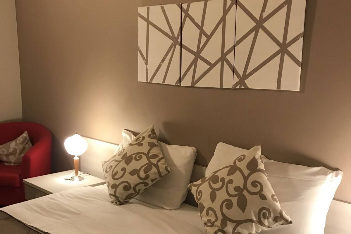 A neatly made bed with patterned pillows, a red chair, and a wall-mounted abstract art piece.