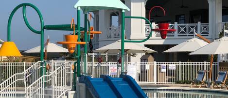 A pool area with a blue slide, a play structure, and umbrellas.