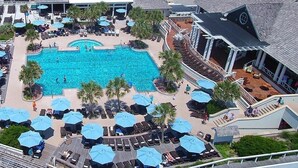 Vista aérea de un resort con una piscina central rodeada de tumbonas y sombrillas, una zona cubierta con asientos y un edificio de techo inclinado.