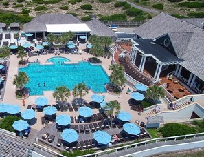 Luchtbeeld van een resort met een centraal gelegen zwembad, omringd door ligbedden en parasols, een overdekt terras met zitplaatsen, en een gebouw met een schuin dak.