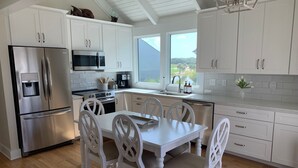 A modern kitchen with white cabinetry, stainless steel appliances, a dining table with chairs, and a view of the outdoors through a large window.