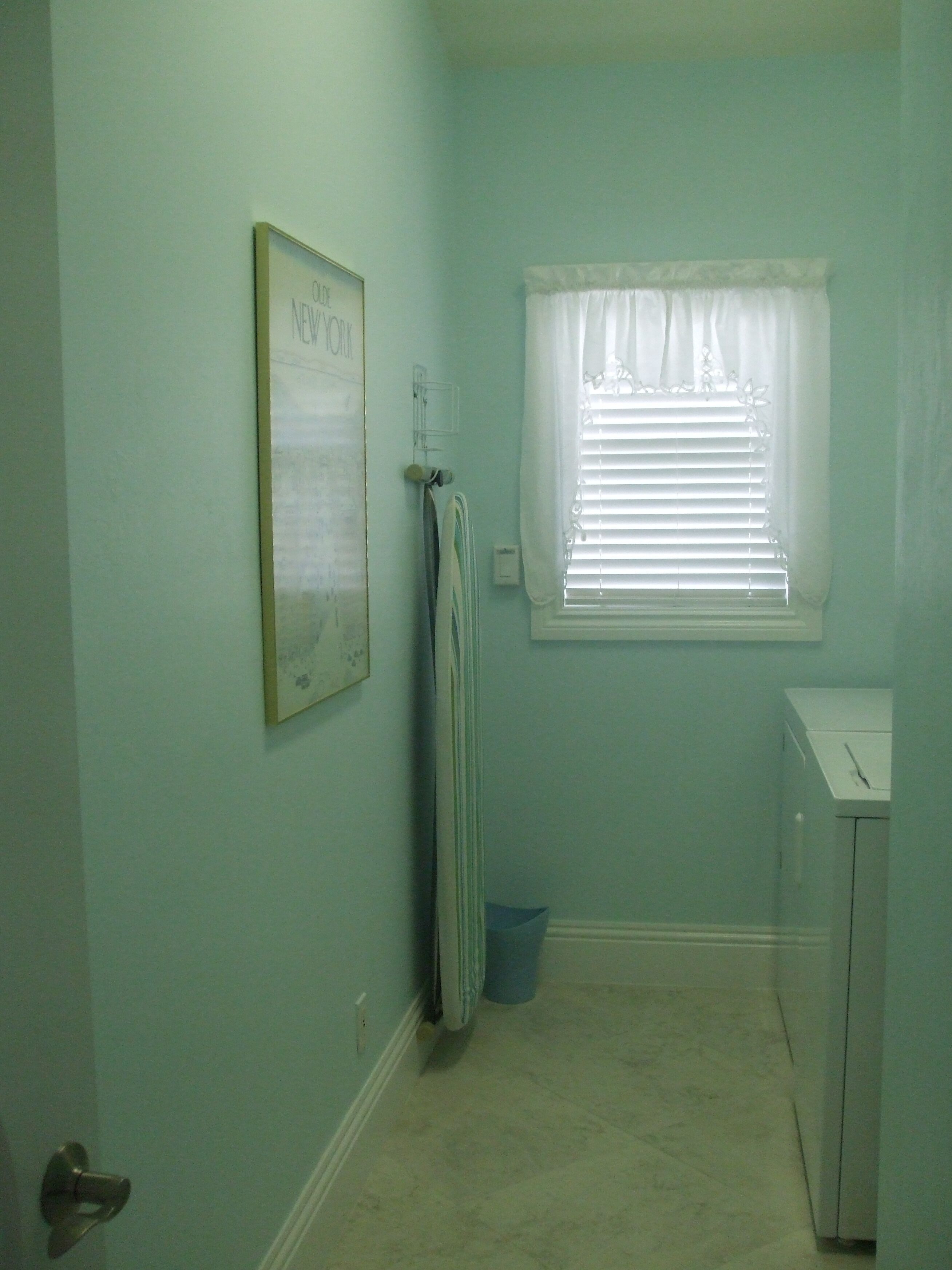 A small, narrow bathroom with light blue walls, a window with white blinds, a framed picture on the wall, and a small cabinet.