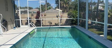 A clear indoor swimming pool with a glass enclosure, surrounded by a patio area with a view of palm trees and houses.