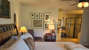 Una camera d'albergo con un letto grande, una scrivania, una sedia, una lampada, quadri appesi al muro e uno spazio doccia.