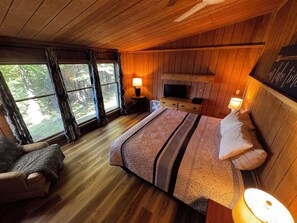 A wooden cabin bedroom with a bed, a chair, a TV, and a window with curtains.