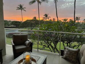 A balcony with a view of a sunset, a chair, a table with a candle, and lush greenery.