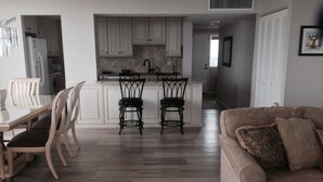 Eine moderne Küche mit weißen Schränken, einer zentralen Kochinsel mit zwei schwarzen Stühlen, einem Esstisch mit Stühlen und einem beigen Sofa mit gemustertem Überwurf.