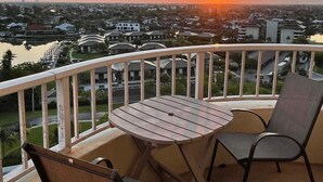 Ein Balkon mit Tisch und zwei Stühlen, mit Blick auf eine Stadtlandschaft im Sonnenuntergang.