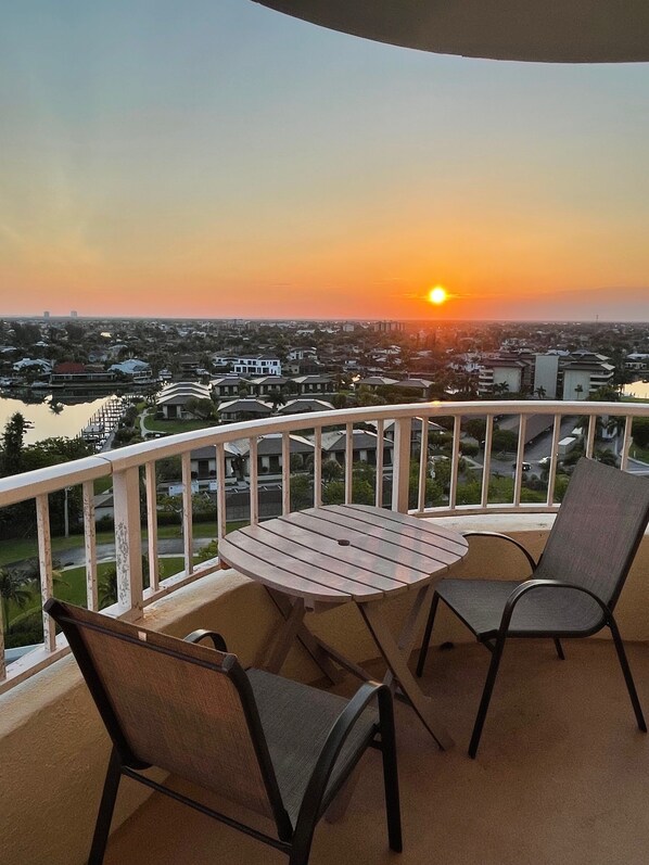 Een balkon met een tafel en twee stoelen, met uitzicht op een stadsgezicht tijdens zonsondergang.