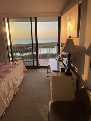 Une chambre d’hôtel équipée d’un lit, d’un bureau, d’une chaise, d’une lampe et offrant une vue sur la mer grâce à de grandes fenêtres.