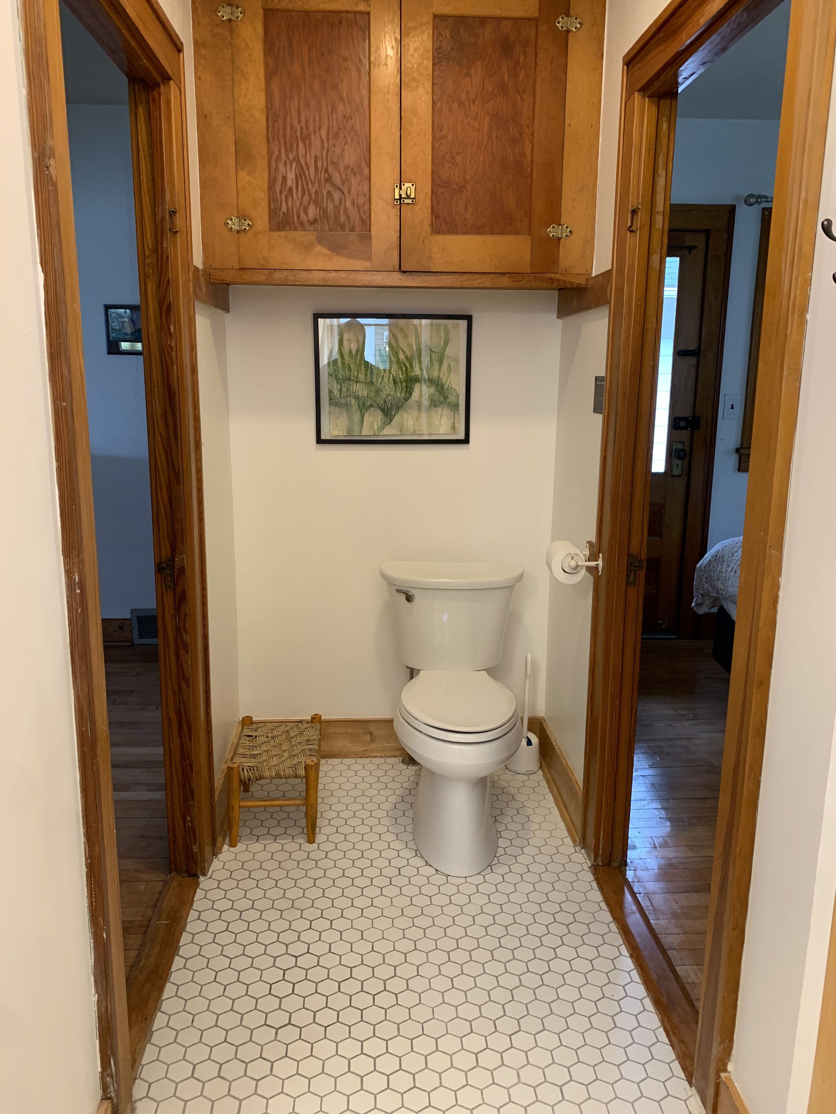 A bathroom with a toilet, wooden cabinets above, a framed picture on the wall, and a small wooden stool.