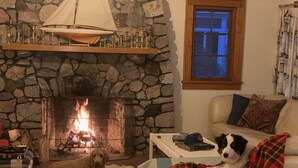 A cozy living room with a stone fireplace, a sofa, a dog, and a window with curtains. - Sandy Beach Cottage on Torch Lake, in Alden (Alden)