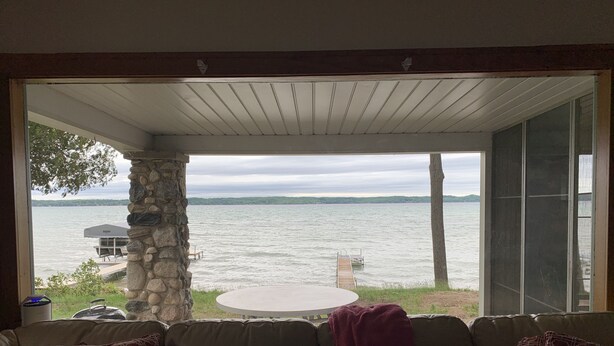 A covered patio with a stone pillar, a round table, and a view of a lake with a dock. - Sandy Beach Cottage on Torch Lake, in Alden (Alden)