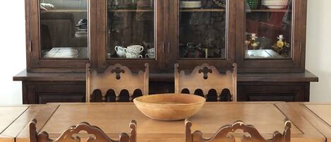 Una mesa de madera para comer con un bol en el centro, rodeada por cuatro sillas con cojines a cuadros. Detrás de la mesa, un armario de madera oscura con puertas de vidrio exhibe diversos utensilios de cocina.