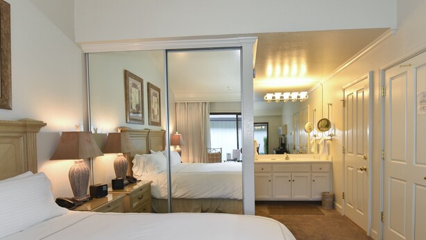 A hotel room with a large bed, bedside lamps, a desk with a lamp, a mirror, a bathroom with a sink and mirror, and a door leading to another room. - Getaway Suite at Silverado in Napa (Napa)