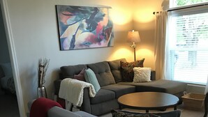 Un soggiorno moderno con un divano grigio, un tavolino rotondo e una panca. Si nota un quadro appeso alla parete, una lampada e una finestra con persiane.