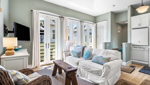 A living room with a white sofa, a wooden coffee table, and a television on a white stand. There is a kitchen area with white cabinets and a refrigerator. - Sandy Souls Cottage & CH - NEWLY RENOVATED! GULF VIEWS! ACCESS TO FITNESS/TENNIS (Rosemary Beach)
