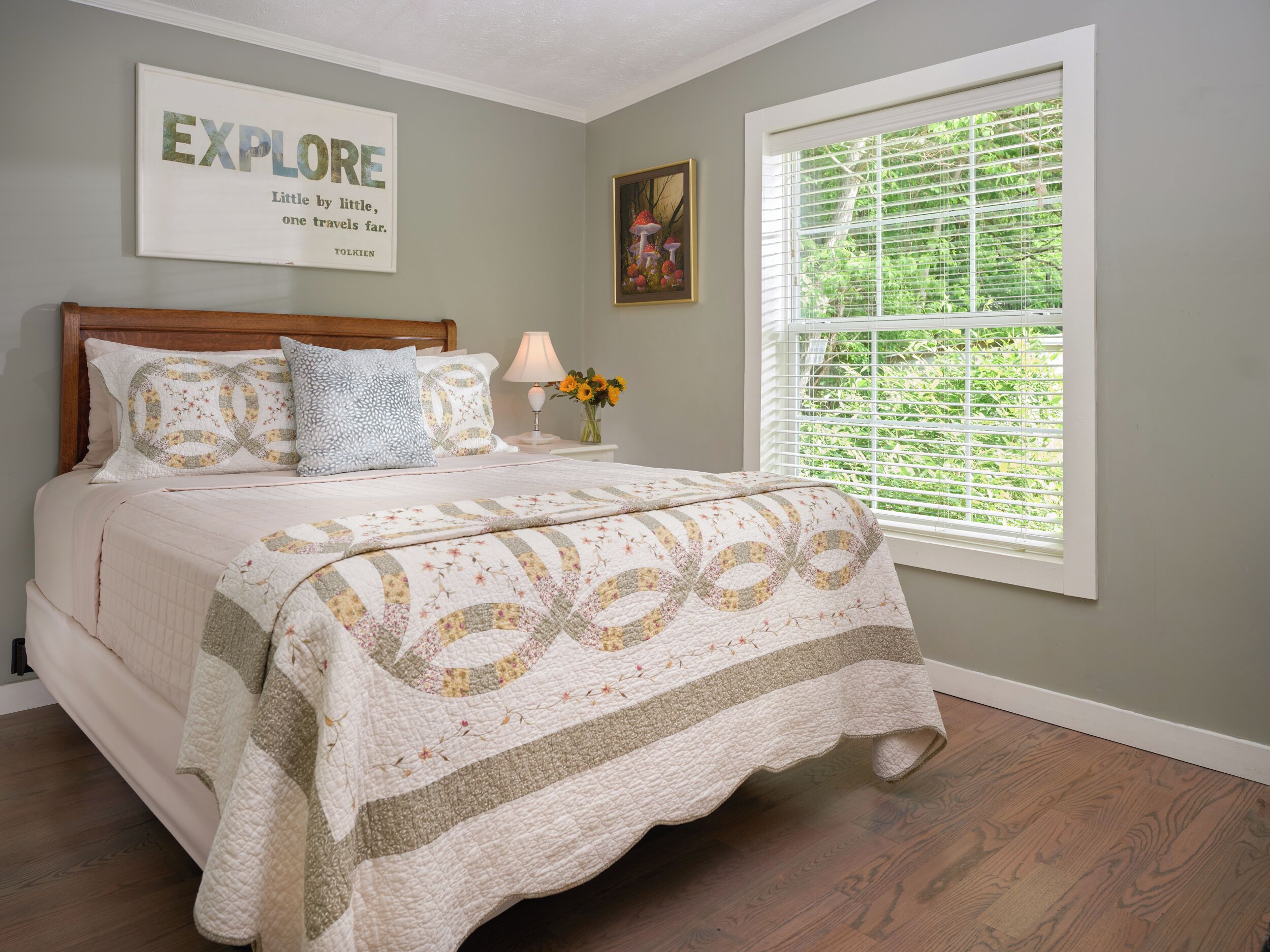 A bedroom with a bed, a nightstand with a lamp, a framed quote on the wall, and a window with blinds.