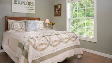 A bedroom with a bed, a nightstand with a lamp, a framed quote on the wall, and a window with blinds.