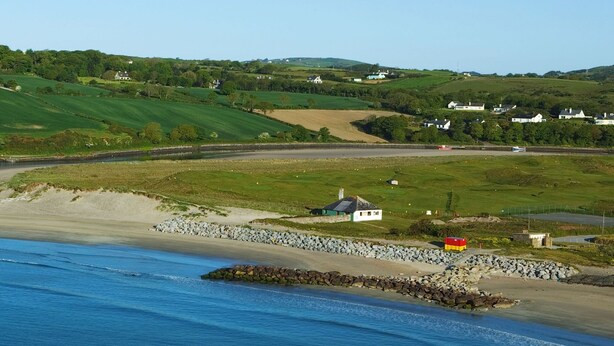 A coastal landscape with a beach, rocky shoreline, and houses in the background.