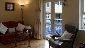 A living room with a chandelier, a red sofa, a grey armchair, a glass coffee table, and a door leading to a balcony.