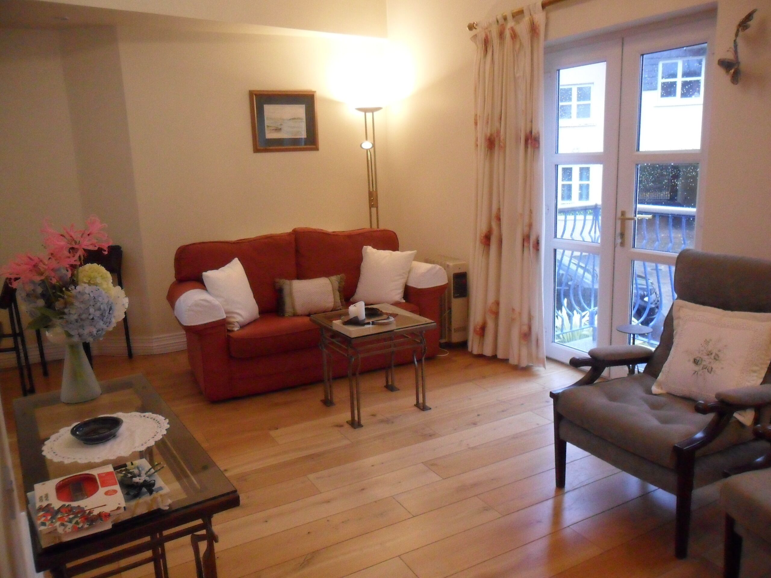 A living room with a red sofa, a glass coffee table, and a grey armchair. There is a vase with flowers on a side table, a lamp, and a framed picture on the wall.