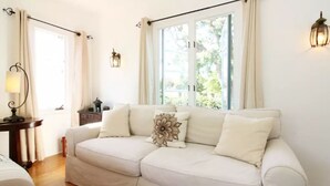 A cozy living room with a beige sofa, a side table with a lamp, and a window with curtains. - Elegant Garden Apartment (Coronado)