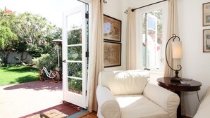 A room with a white sofa, a small table with a lamp, and a door leading to a garden. - Elegant Garden Apartment (Coronado)